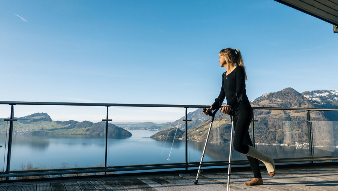 Eine Frau mit eingegipstem Bein steht mit Krücken auf einer Terrasse mit Blick auf eine Seenlandschaft mit bergigem Umland.