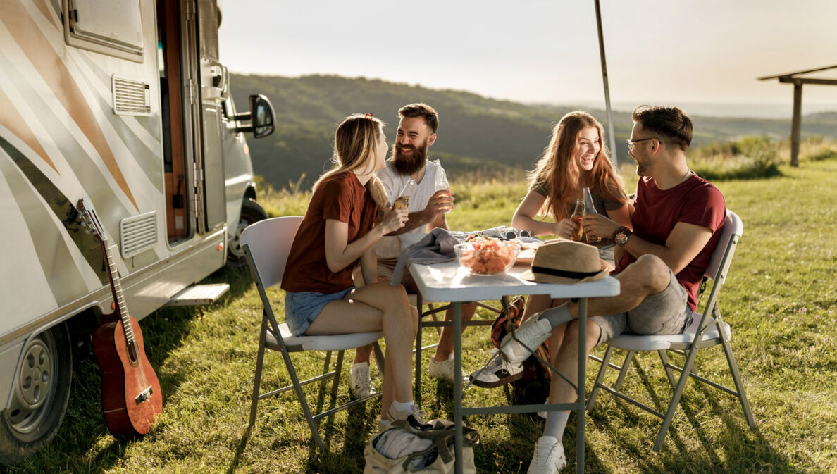 Vier gut gelaunte Personen in Sommerkleidung sitzen an einem Campingtisch neben einem Wohnmobil auf einer Rasenfläche vor Hügellandschaft.