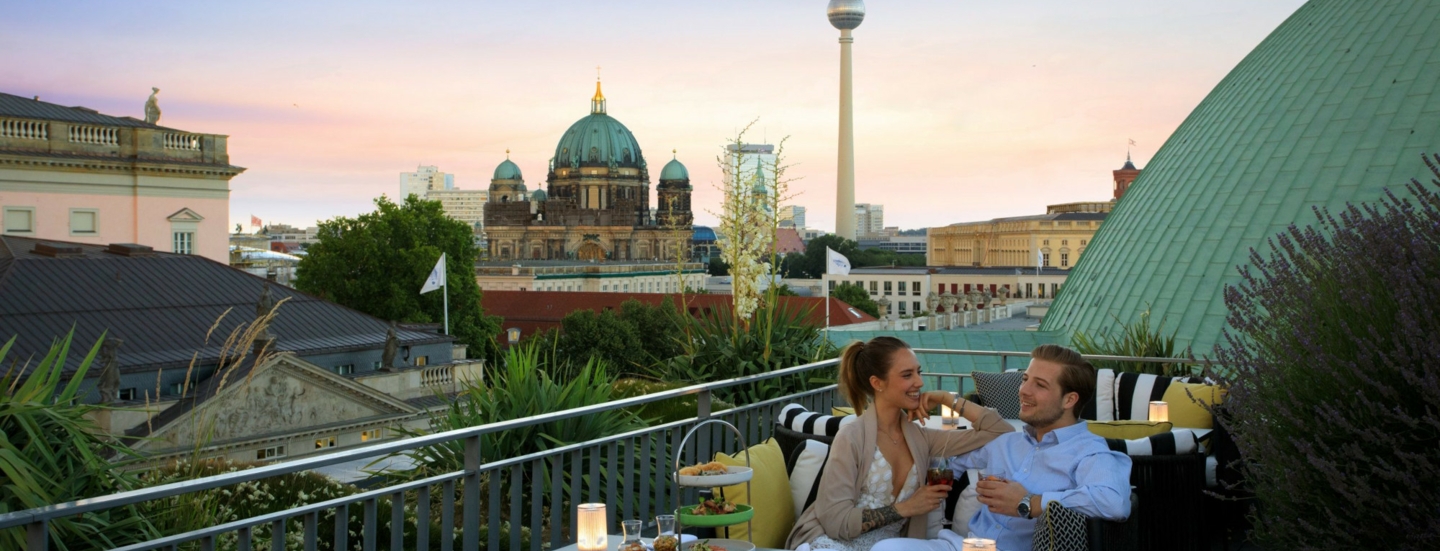 Ein junges Paar sitzt gut gelaunt an einem Tisch auf einer Dachterrasse, im Hintergrund Berliner Dom und Fernsehturm.