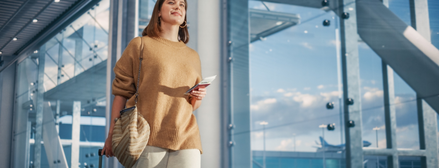 eine Frau am Flughafen, die mit einer Hand einen Koffergriff festhält und in der anderen Papiere hält.