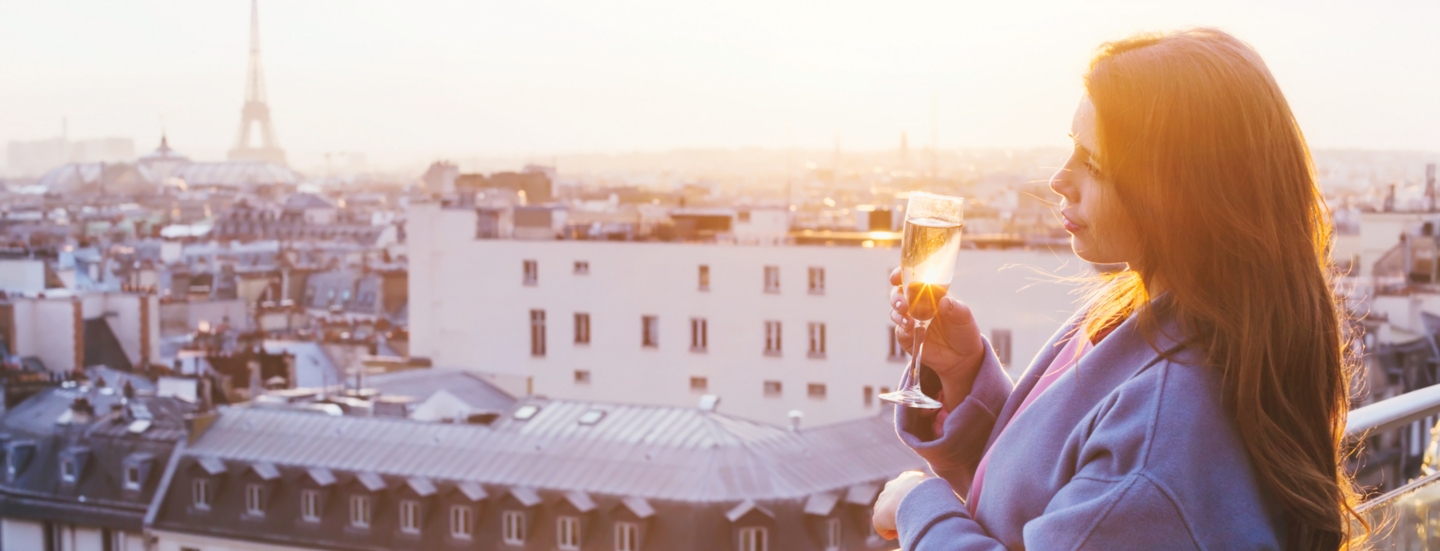 Eine Frau, die bei Sonnenuntergang in Paris auf einem Balkon steht und ein Glas Sekt in der Hand hält.