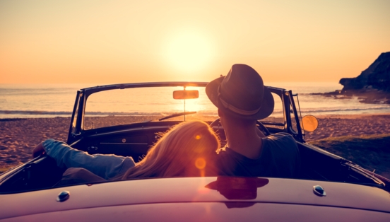 Rückansicht eines Paares in einem Cabriolet an einem Sandstrand bei Sonnenuntergang, sie hat den Kopf auf seine Schulter gelehnt.