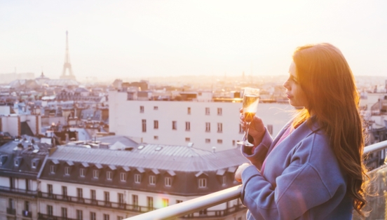 Eine Frau, die bei Sonnenuntergang in Paris auf einem Balkon steht und ein Glas Sekt in der Hand hält.