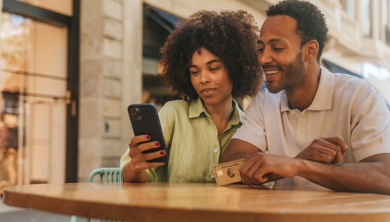 Ein Paar steht an einem Tisch auf einer Einkaufsstraße und schaut gemeinsam auf ein Smartphone, er hält eine goldene Kreditkarte von American Express in der Hand.