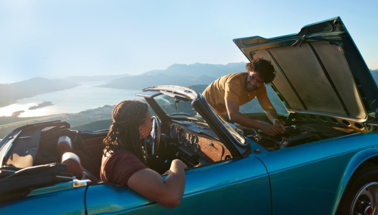 Ein Paar in einem liegengebliebenen Cabriolet an einer Meeresküste bei Sonnenschein, er kontrolliert den Motor, sie sitzt auf dem Beifahrersitz.