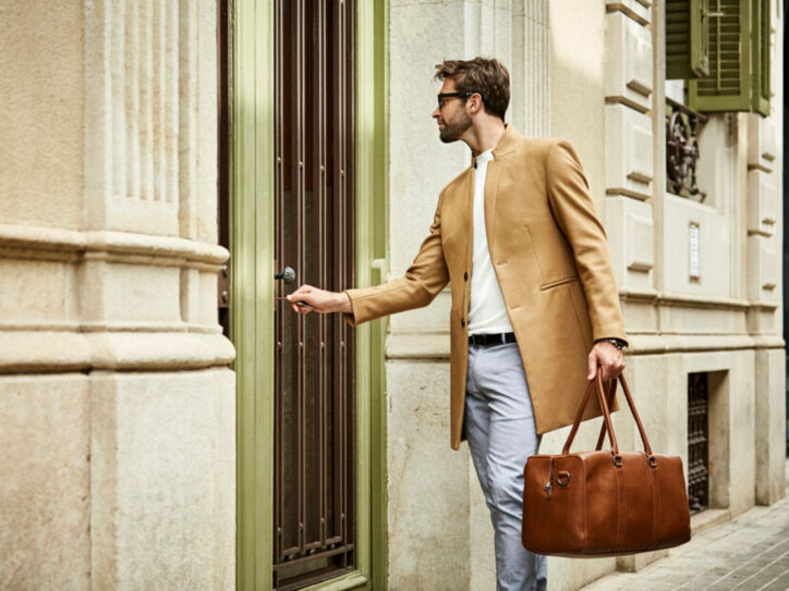 Ein junger, moderner Mann mit Sonnenbrille, beigem Wollmantel und brauner Ledertasche schließt eine Haustür auf