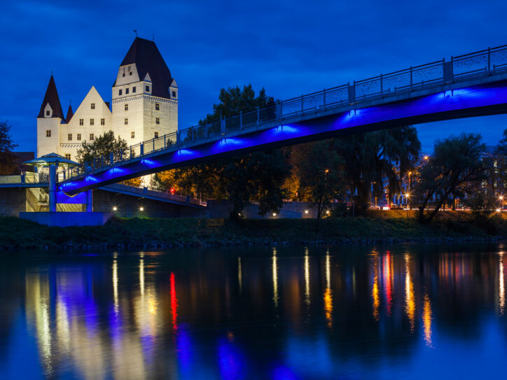 Das beleuchtete Neue Schloss in Ingolstadt bei Nacht, im Vordergrund führt eine Brücke über die Donau