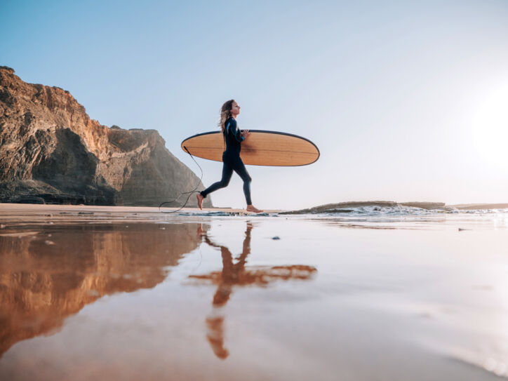 Eine Frau im Neoprenanzug trägt ihr Surfbrett unterm Arm ins Meer