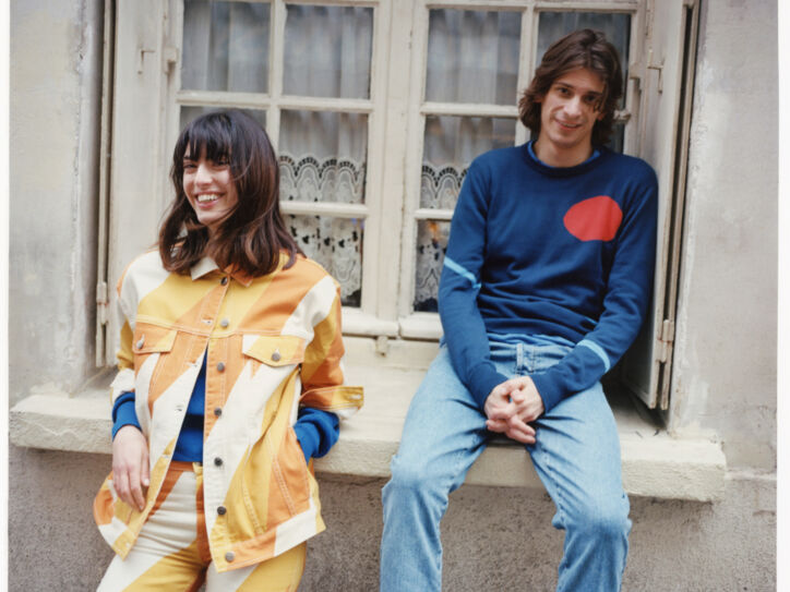 Ein Mann und eine Frau vor einem Fenster eines alten Gebäudes tragen farbige und lässige Outfits.