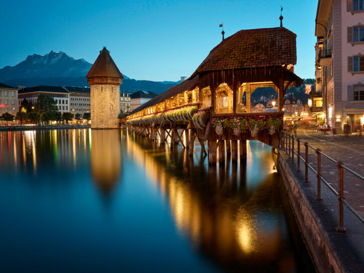 Eine überdachte und beleuchtete Holzbrücke über einem Fluss, ein Wasserturm und eine Stadt und Berge im Hintergrund.