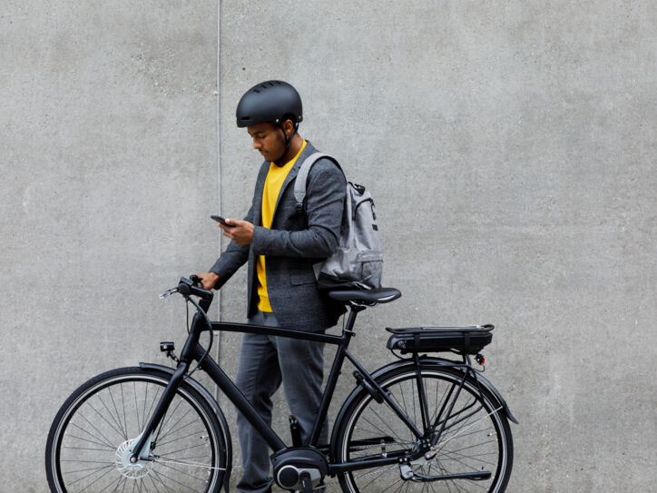 Ein junger Mann mit Rucksack und Fahrradhelm steht neben seinem E-Bike und schaut auf sein Smartphone.