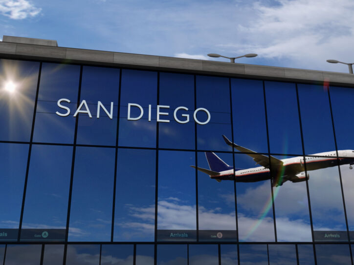 Flugzeug beim Abflug, das sich in den verspiegelten Fenstern einer Flughafenhalle spiegelt, auf denen die Aufschrift „San Diego“ zu lesen ist.