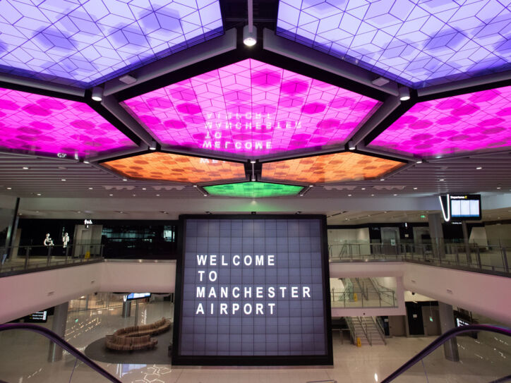 Menschenleere Flughafenhalle mit einem Bildschirm im Mittelpunkt, auf dem „Welcome to Manchester Airport“ steht und bunt erleuchteten LED-panels an der Decke.