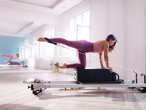 Eine Frau in pinker Sportkleidung kniet auf einer Box auf einem Pilates Reformer in einem hellen Raum.