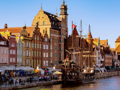 Personen an einer Uferpromenade mit den mittelalterlichen Handelshäusern und historischem Mastschiff.