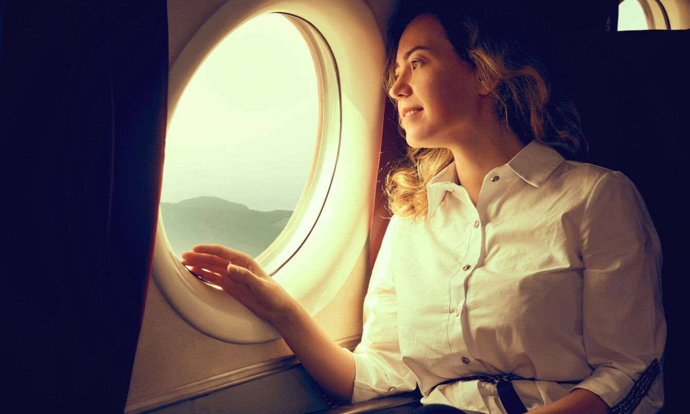 Eine junge Frau in weißer Bluse schaut aus dem Fenster eines Flugzeuges auf eine Landschaft.