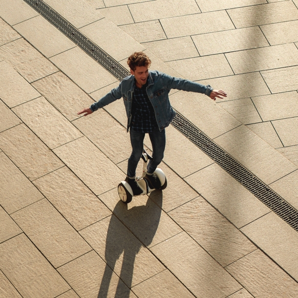 Ein lachender, junger Mann in Jeansjacke auf einem weißen Hoverboard