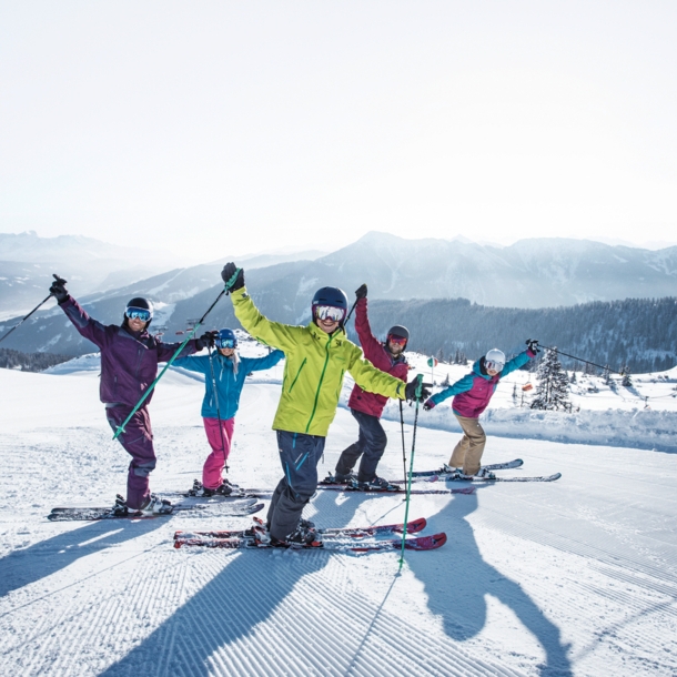 Fünf Skifahrende in bunter Kleidung winken in die Kamera auf einer Piste vor winterlichem Bergpanorama