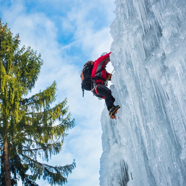 Ein Mensch klettert an einem gefrorenen Wasserfall hinauf, daneben eine große Tanne.