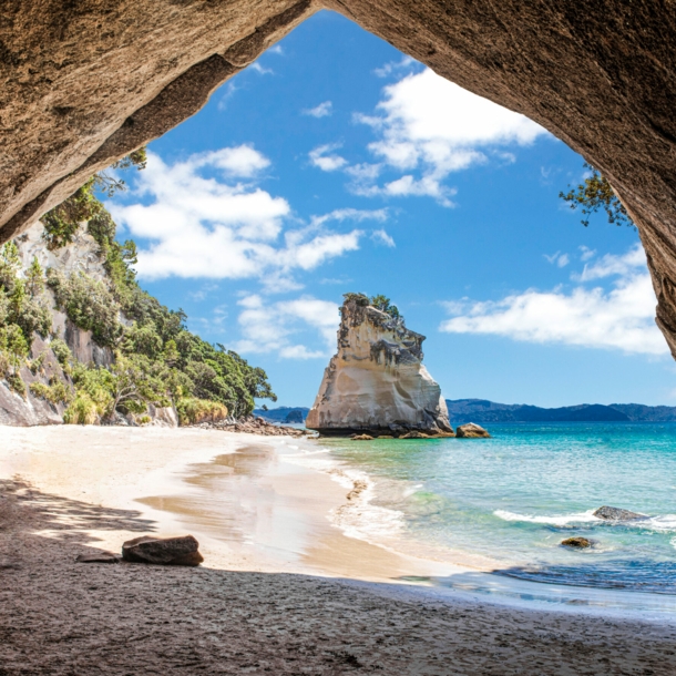 Im Vordergrund ein Felsbrocken, dahinter ein Sandstrand, umringt von Felsen und türkisfarbenem Meer