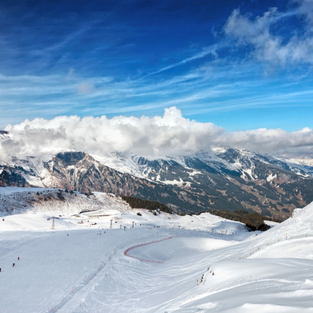 Blick von oben auf eine Piste mit Skifahrer:innen, im Hintergrund Berge