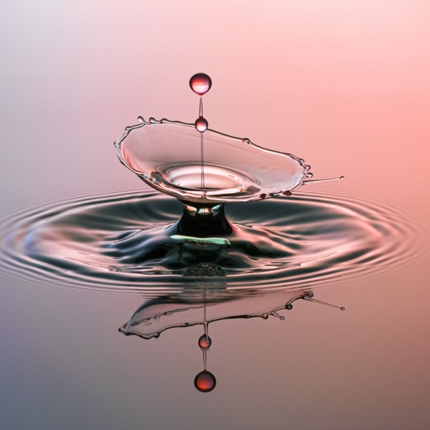 Eine lilafarben und rot angestrahlte Wasserfläche, in die ein Wassertropfen fällt.