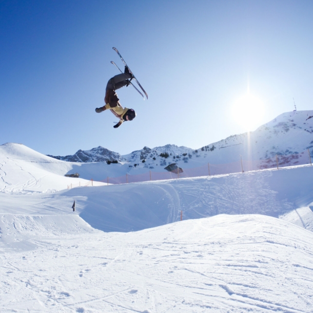 Eine Person, die mit Ski einen Salto in der Luft macht, im Hintergrund ein schneebedeckter Berg