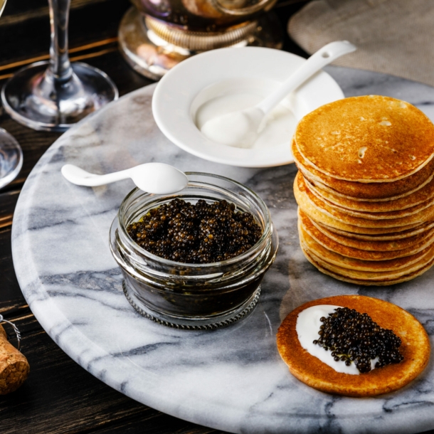 Kaviar im Glas, ein Stapel Blinis und Löffel auf einem Marmortisch, daneben Sektkorken und -gläser