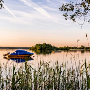 Ein See mit einem Boot und einem Sprungturm in der Abendsonne, im Vordergrund Schilf