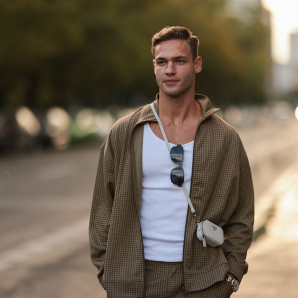 Ein Mann trägt einen braunen Trainingsanzug, hat eine Sonnenbrille am Shirt und eine kleine Tasche umgehängt.