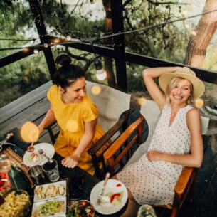 Zwei Frauen sitzen gut gelaunt beim sommerlichen Outdoor-Dinner an einem Tisch auf einer Holzterrasse