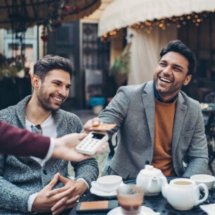 Ein junger, gut gelaunter Mann bezahlt kontaktlos mit seiner American Express Gold Card im Beisein seiner Freunde an einem Tisch in einem Café 