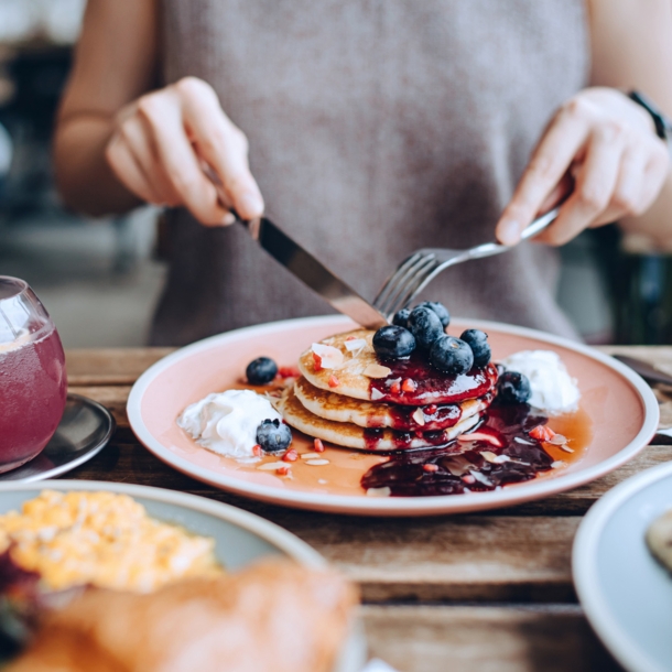 Eine Person sitzt an einem gedeckten Tisch vor einem Teller mit Pancakes im Außenbereich eines Cafés