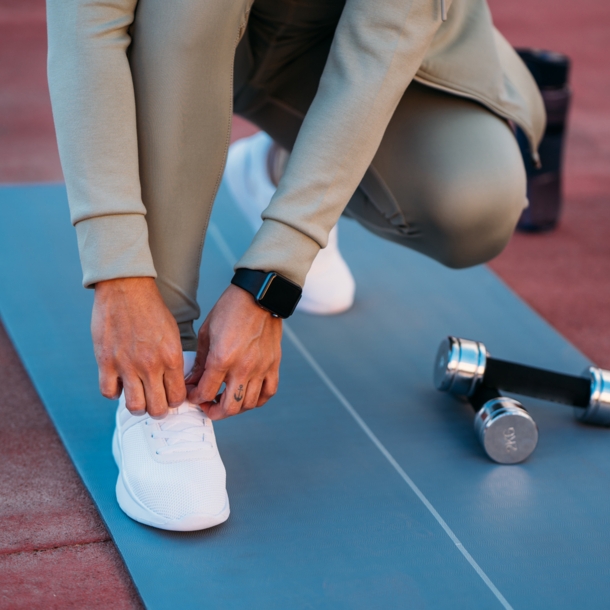 Eine Person in Sportkleidung mit Fitnessuhr am Handgelenk bindet sich die Schnürsenkel auf einer Sportmatte.