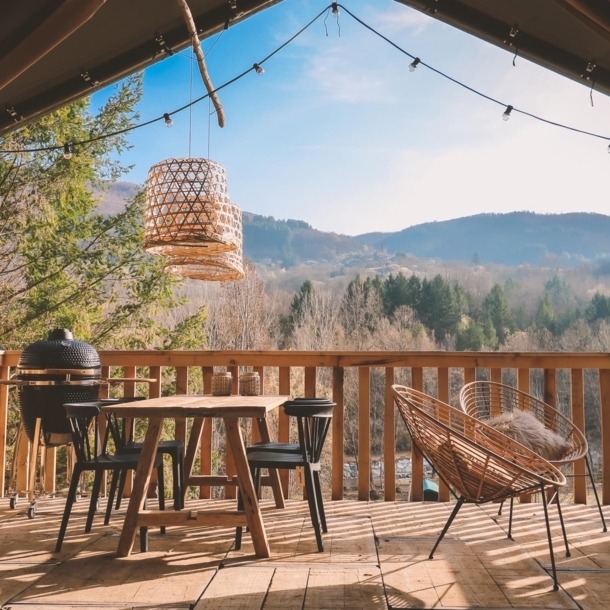 Holzterrasse mit Tisch, Stühlen und Lampen, im Hintergrund bewaldete Hügel