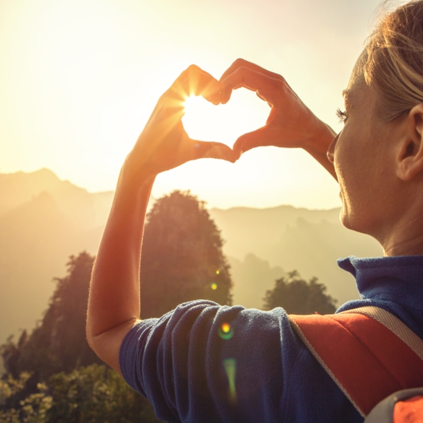 Eine junge Frau blickt in die Natur und formt ihre Hände zu einem Herz, durch das die Sonne scheint