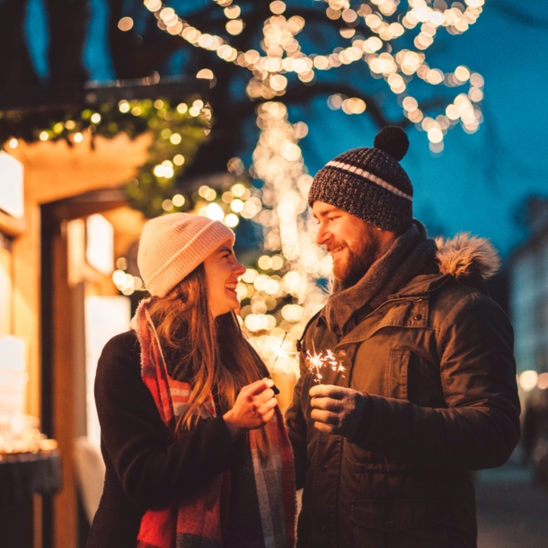 Eine Frau und ein Mann stehen auf dem Weihnachtsmarkt und schauen sich an, in ihren Händen halten sie Wunderkerzen