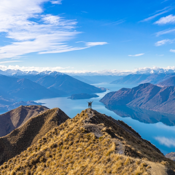 Ein Paar steht auf einem Gipfel vor Seenlandschaft und Bergpanorama der Neuseeländischen Alpen