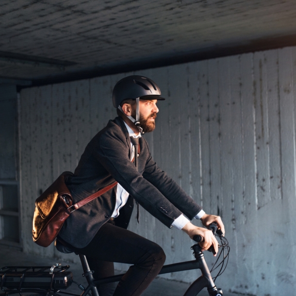 Seitenansicht einer Person im Anzug, mit Umhängetasche und Helm, die auf einem Fahrrad fährt