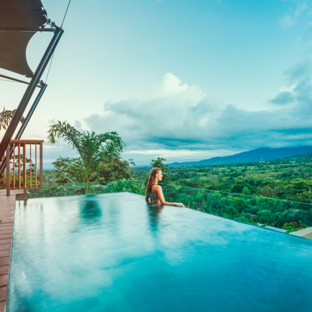 Profilansicht einer Frau, die aus einem Infinity-Pool über eine tropische Hügellandschaft schaut