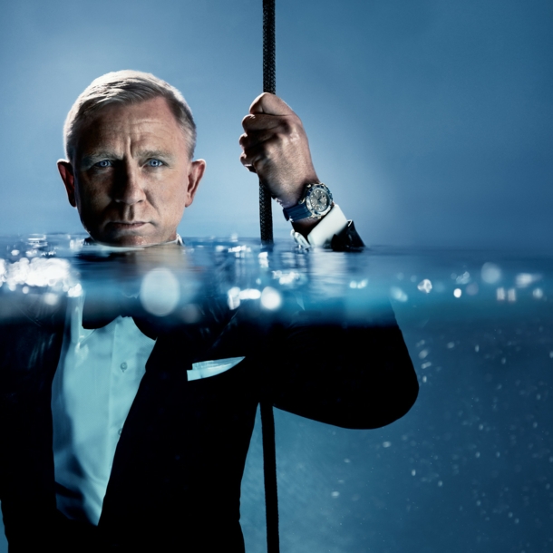 Porträt von Daniel Craig im schwarzen Smoking im Wasser, das ihm bis zum Hals steht; an seiner hochgehaltenen Hand trägt er eine Taucheruhr der Marke OMEGA