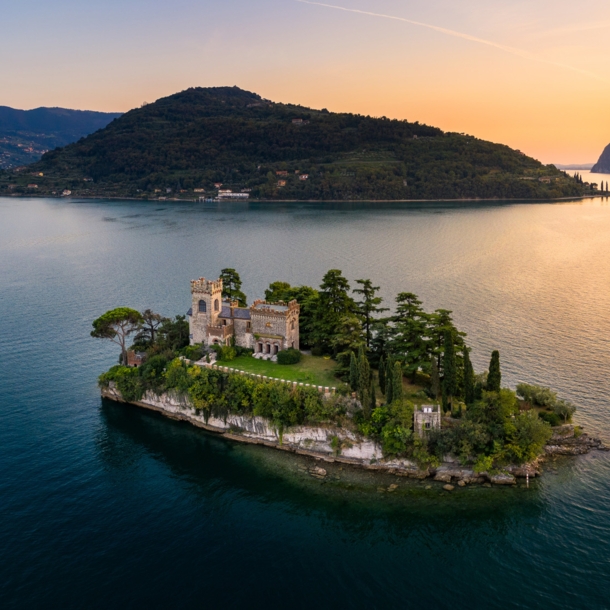 Luftaufnahme einer kleinen Felsinsel mit Burg und Bäumen in einem großen See bei Sonnenuntergang