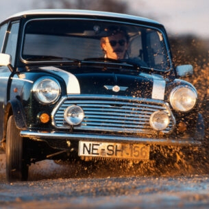 Ein Mann fährt in einem schwarzen MINI Cooper 1.3 mit Rallystreifen durch eine Schlammpfütze