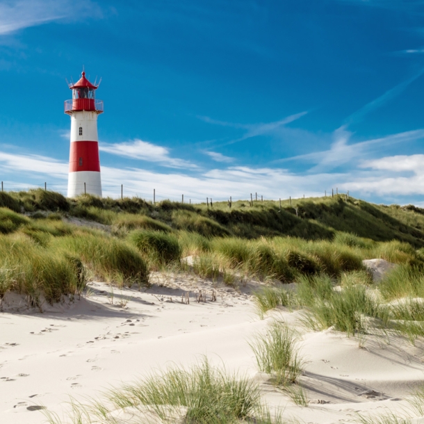 Ein rot-weiß geringelter Leuchtturm in einer grasbewachsenen Dünenlandschaft vor blauem Himmel