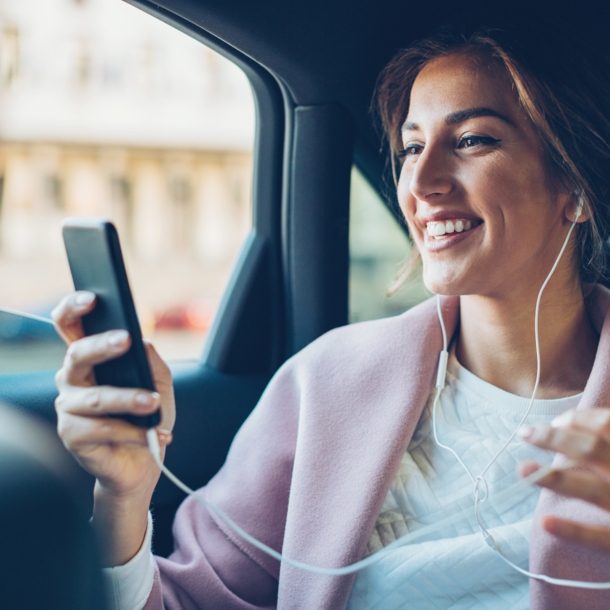 Eine junge Frau mit Kopfhörern auf dem Rücksitz eines Autos schaut lachend auf das Display ihres Smartphones.