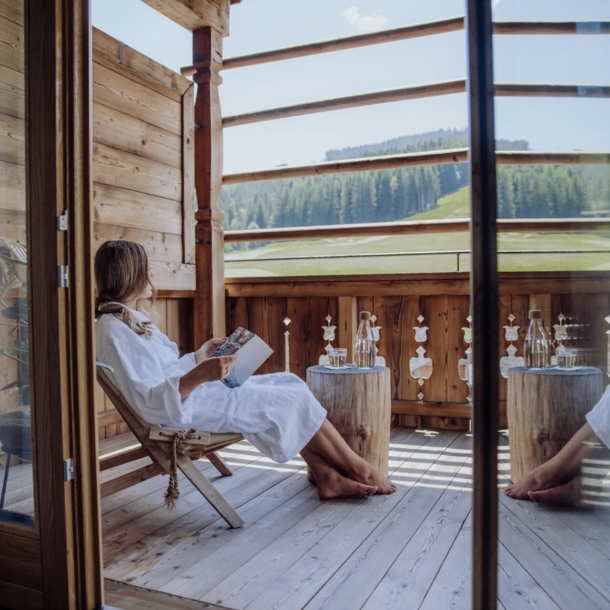 Eine Frau in weißem Bademantel sitzt mit einem Buch auf einer Terrasse eines Holzchalets mit Blick in die Berge