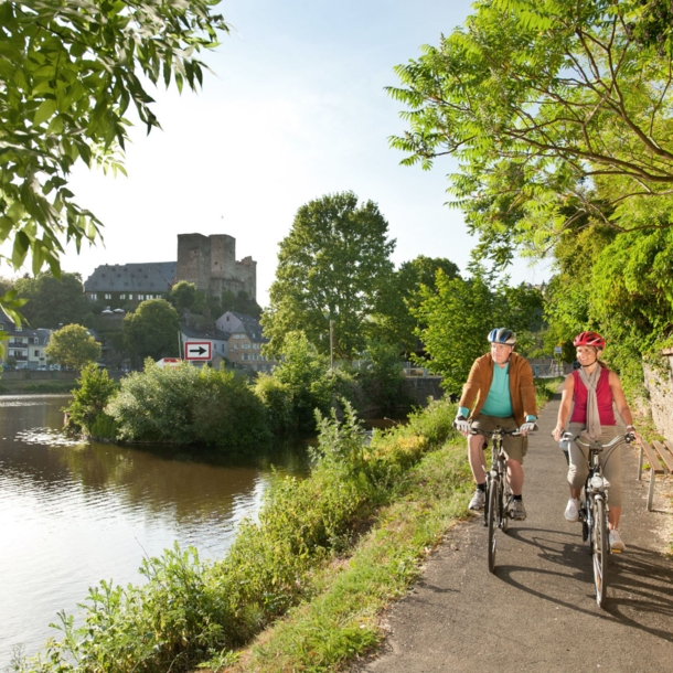 Ein älteres Paar fährt auf Fahrrädern auf einem asphaltierten Radweg entlang eines Flusses, im Hintergrund eine mittelalterliche Burgruine in einer Ortschaft