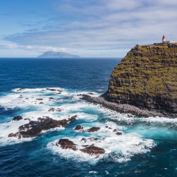 Luftaufnahme einer Steilklippe mit Leuchtturm und der Küste vorgelagerte Felsen im Meer, im Hintergrund eine Insel