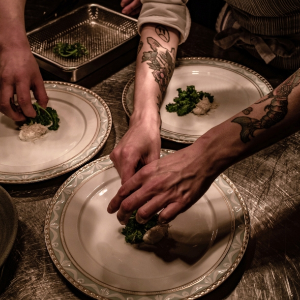 Nahaufnahme von Händen beim Anrichten von Speisen auf großen, weißen Tellern in einer Restaurantküche.