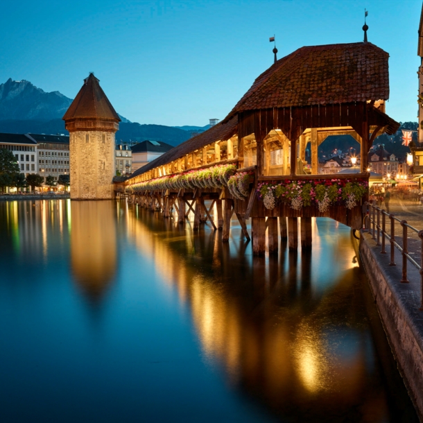 Eine überdachte und beleuchtete Holzbrücke über einem Fluss, ein Wasserturm und eine Stadt und Berge im Hintergrund.